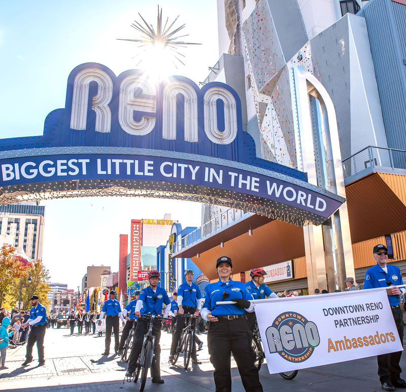 Ambassadors walking and riding their bikes holding up a sign that says "Downtown Reno Partnership Ambassadors" in front of the Reno Arch.