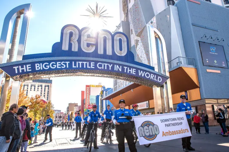 Ambassadors walking and riding their bikes holding up a sign that says "Downtown Reno Partnership Ambassadors" in front of the Reno Arch.